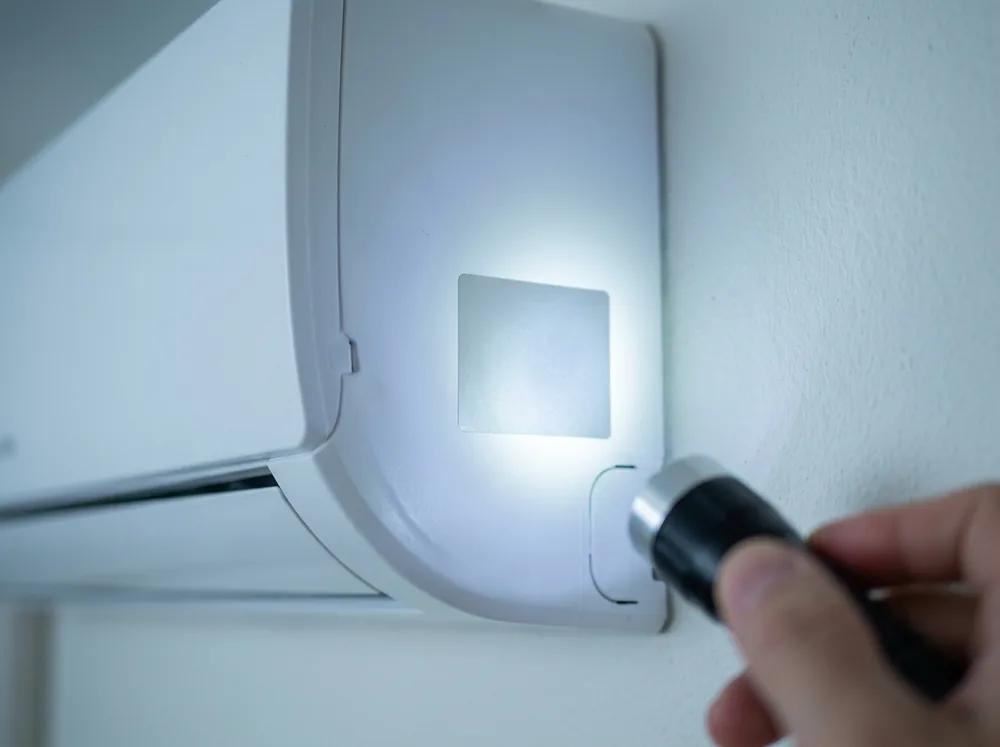 イメージ:エアコン室内機の側面に貼られた型式表示ラベルを、懐中電灯で照らして確かめている手元
